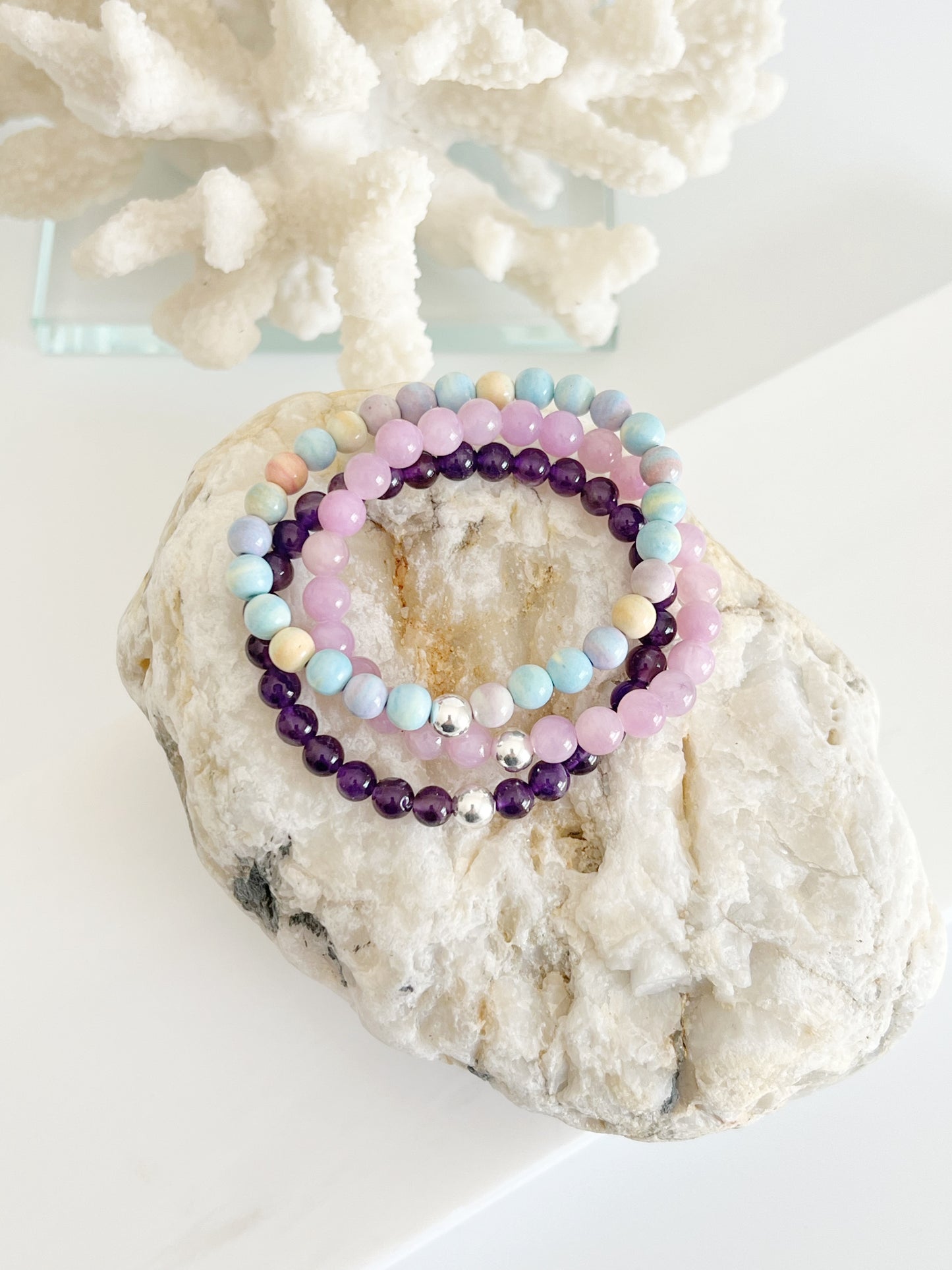 three stretch bracelets on top of each other, amethyst (deep purple ) a candy colored bracelet ( yellow, lilac, blue, green ) and a kunzite ( lilac ) on top of whitish and light brown large rock in front of white coral