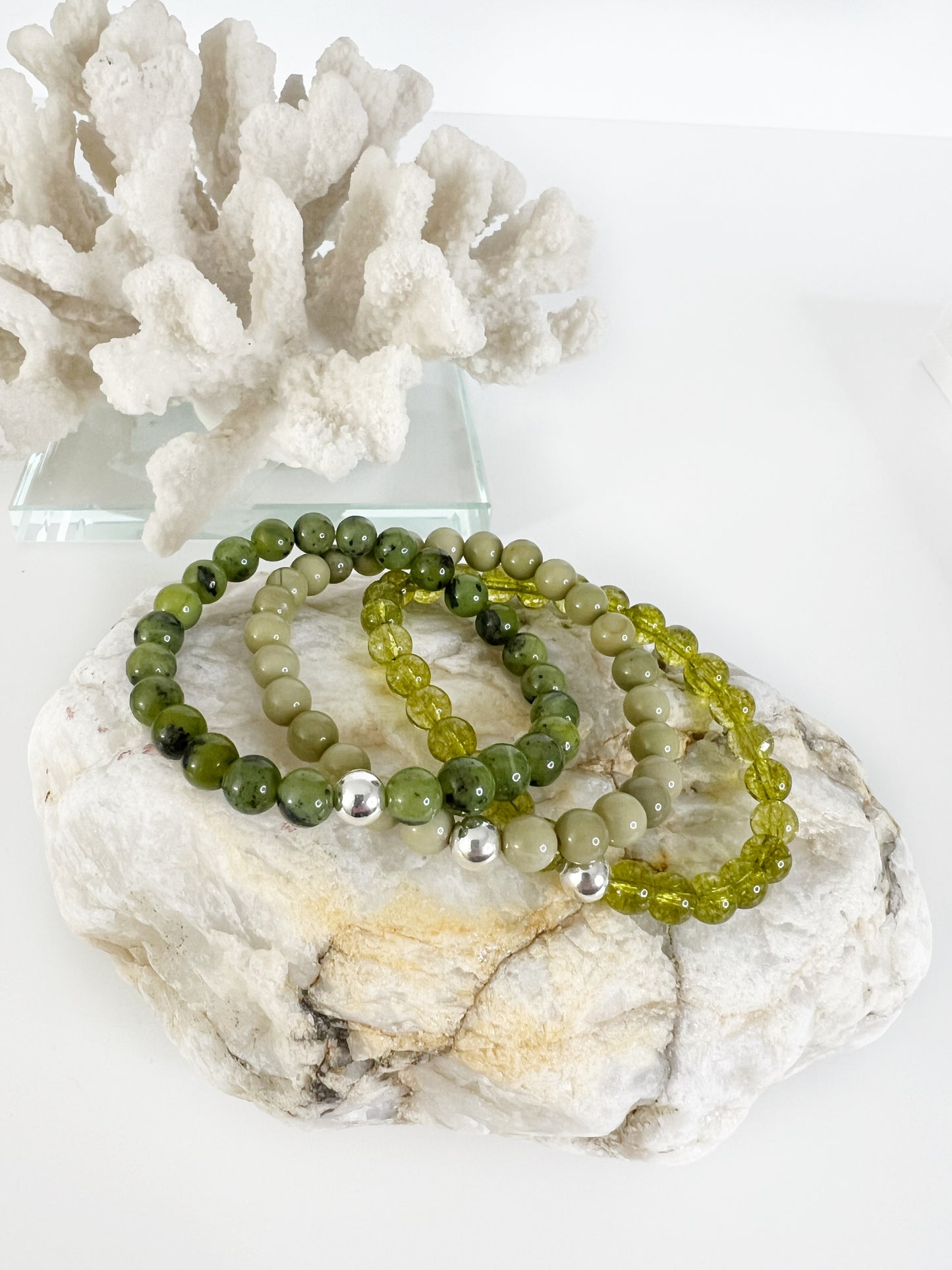 three different green stretch bracelets with one silver bead, on top of a brownish rock with white coral behind it.