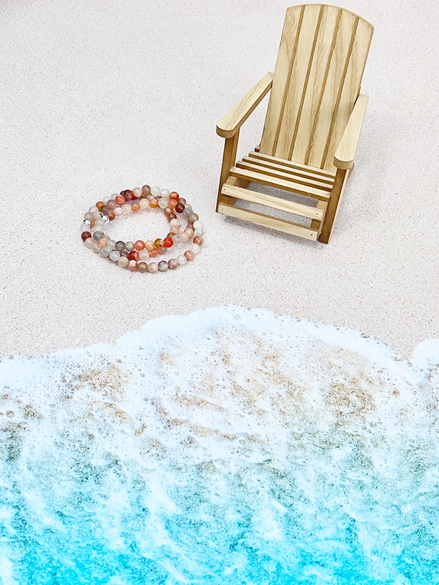 peach, grey and white moonstone stretch bracelet w/ one sterling silver connector bead on the beach next to a wooden adirondak chair