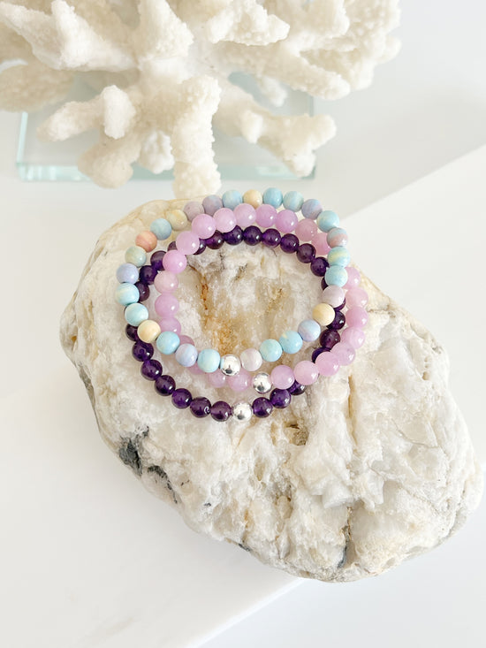 three stretch bracelets on top of each other, amethyst (deep purple ) a candy colored bracelet ( yellow, lilac, blue, green ) and a kunzite ( lilac ) on top of whitish and light brown large rock in front of white coral 