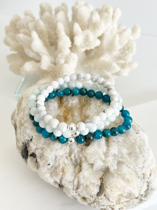 Three stretch bracelets stacked on top of each other. One is light green, one white and one mixed turquoise blue. They each have one silver bead and they on top of a light brown rock with white coral behind them.