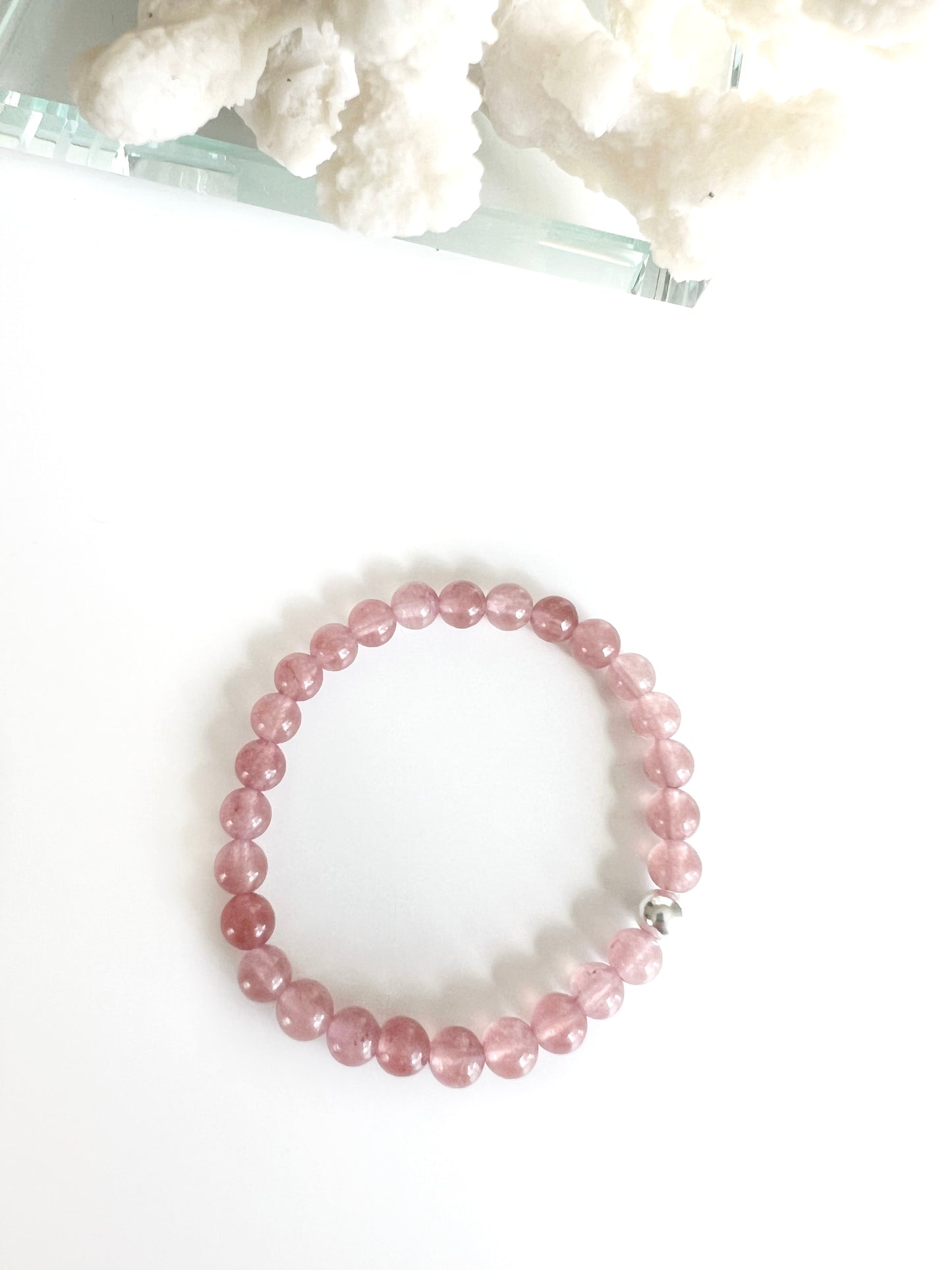 Strawberry Quartz stretch bracelet with one silver bead. Each bead is dark translucent pink. There's part of a white coral behind it.