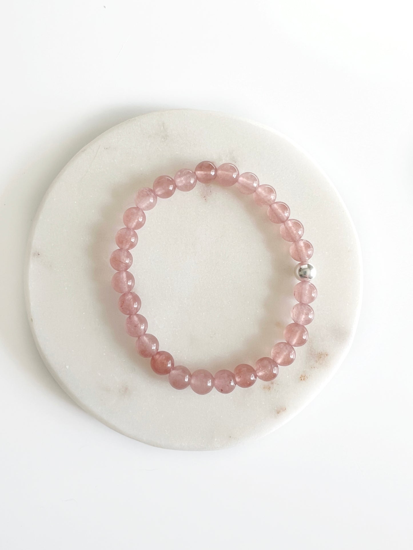 Strawberry Quartz stretch bracelet with one silver bead. Each bead is dark translucent pink. It's sitting on a small flat white dish.