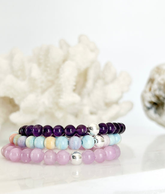 three stretch bracelets on top of each other, amethyst (deep purple ) a candy colored bracelet ( yellow, lilac, blue, green ) and a kunzite ( lilac ) in front of white coral and small part of the a whitish rock on the right side