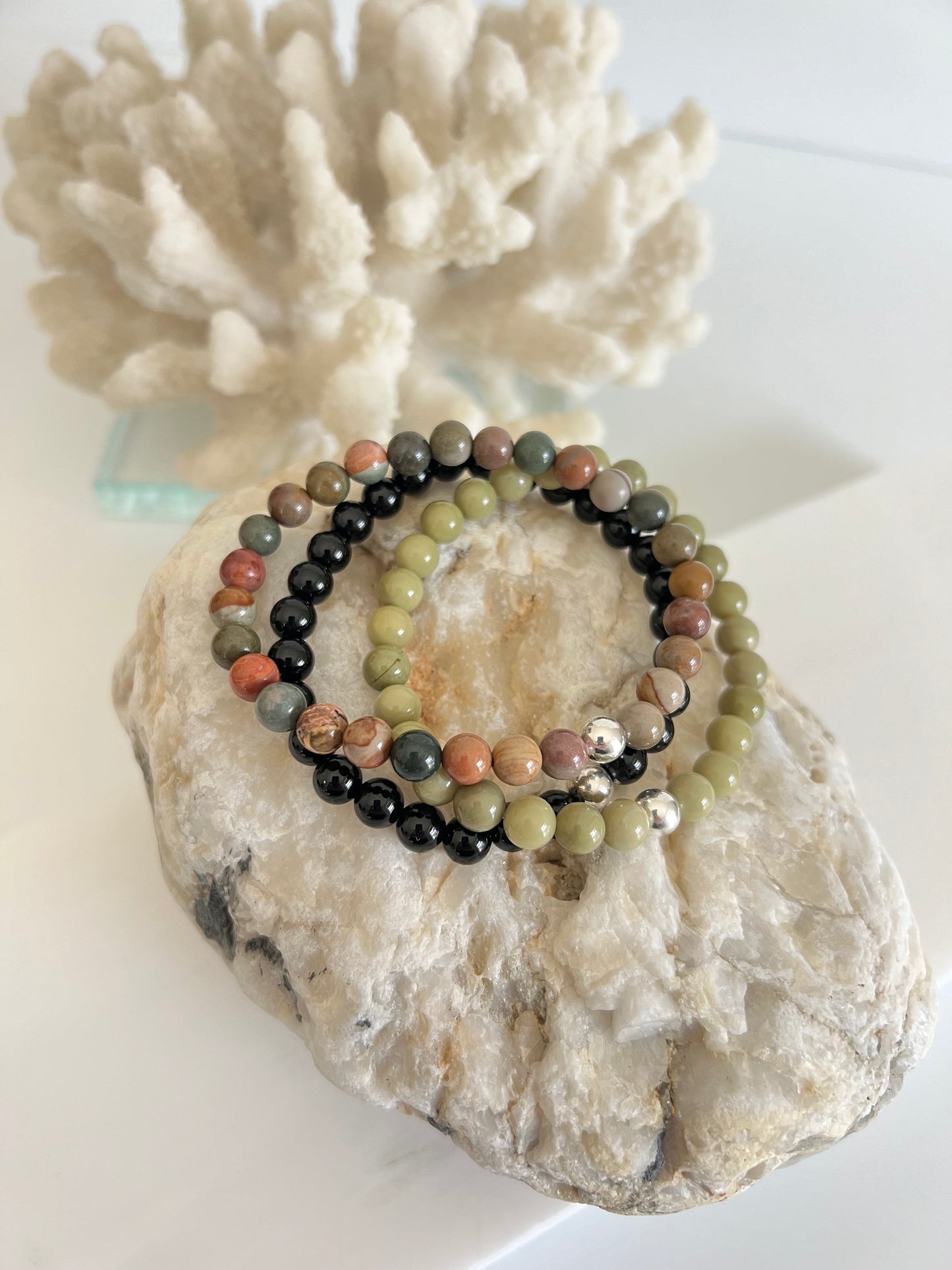 three stretch bracelets on top of a whitish and brown rock with a white piece of coral behind it. the bracelets are multi brown, red and green colored on the top, then avocado green in the middle and a black one on the bottom