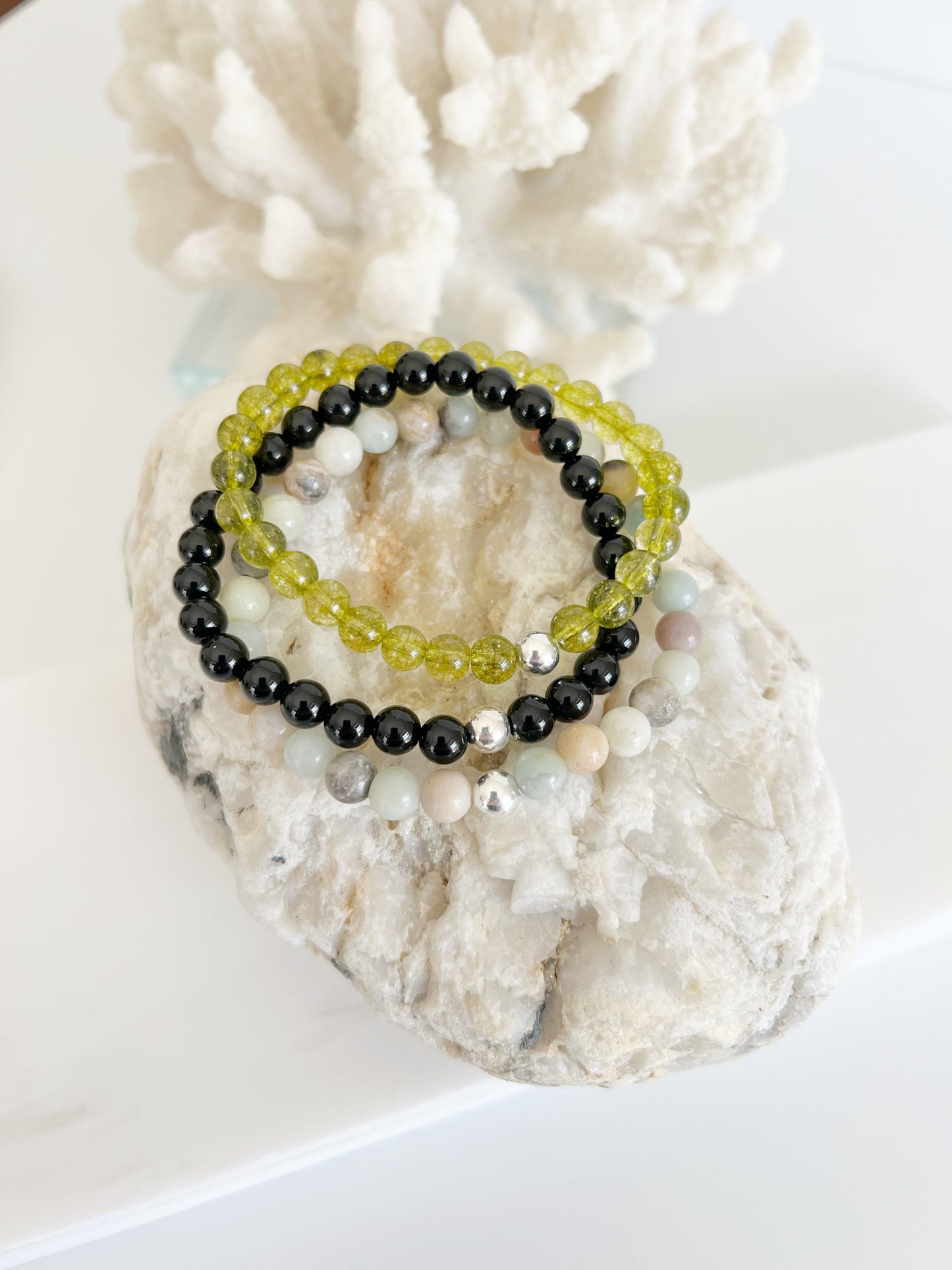 three stretch bracelets stacked on top of each other on a light brown rock white coral behind it.  One transluscent green one, a multi colored one with light blue, yellow, black and white and a black one. All have one silver bead.