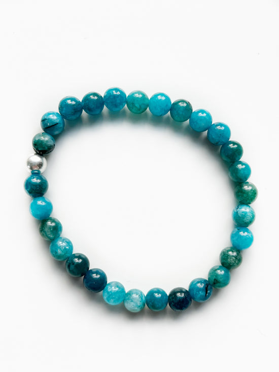 A picture of a stretch bracelet on a white background. The stretch bracelet is all Apatite gemstone beads which are shades of light to dark teal like ocean waves. There is one Sterling Silver bead on the bracelet