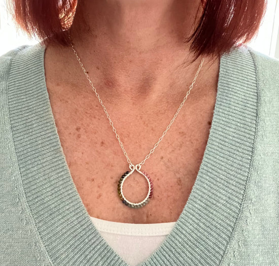 A woman wearing a sage green v neck sweater with a white tank top underneath wearing a silver round pendant necklace with tiny tourmaline beads wire wrapped around it.
