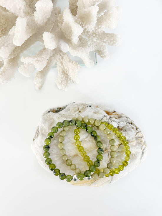 three different green stretch bracelets with one silver bead, on top of a brownish rock with white coral behind it.