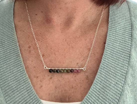 A woman wearing a sage green v neck sweater with a small bit of white tank top showing underneath, she's wearing a sterling silver bar necklace with 9 round Tourmaline beads in colors from black to green to pink wire wrapped to the bottom of the necklace.