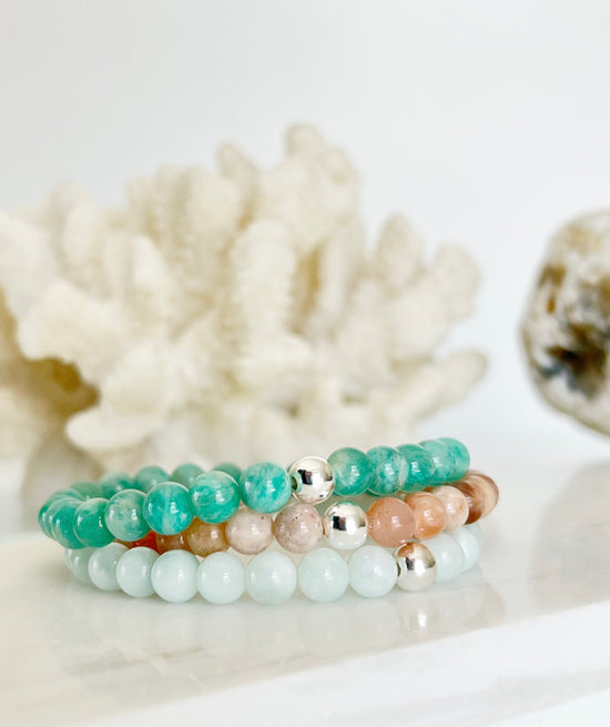 Modern everyday gemstone stretch bracelet trio of Amazonite, Peach Moonstone and Green Moonstone with white coral and a brownish rock behind them.