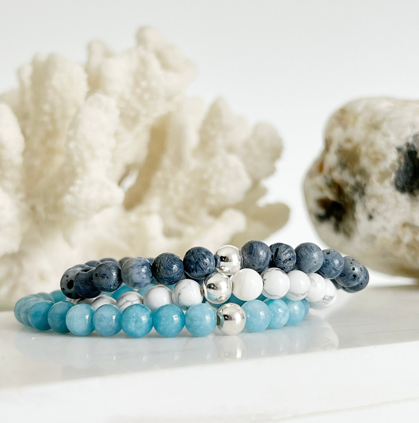 Modern everyday gemstone stretch bracelet trio of Blue Coral, Howlite and Blue Chalcedony stacked on top of each other with white coral and brownish rock behind them.