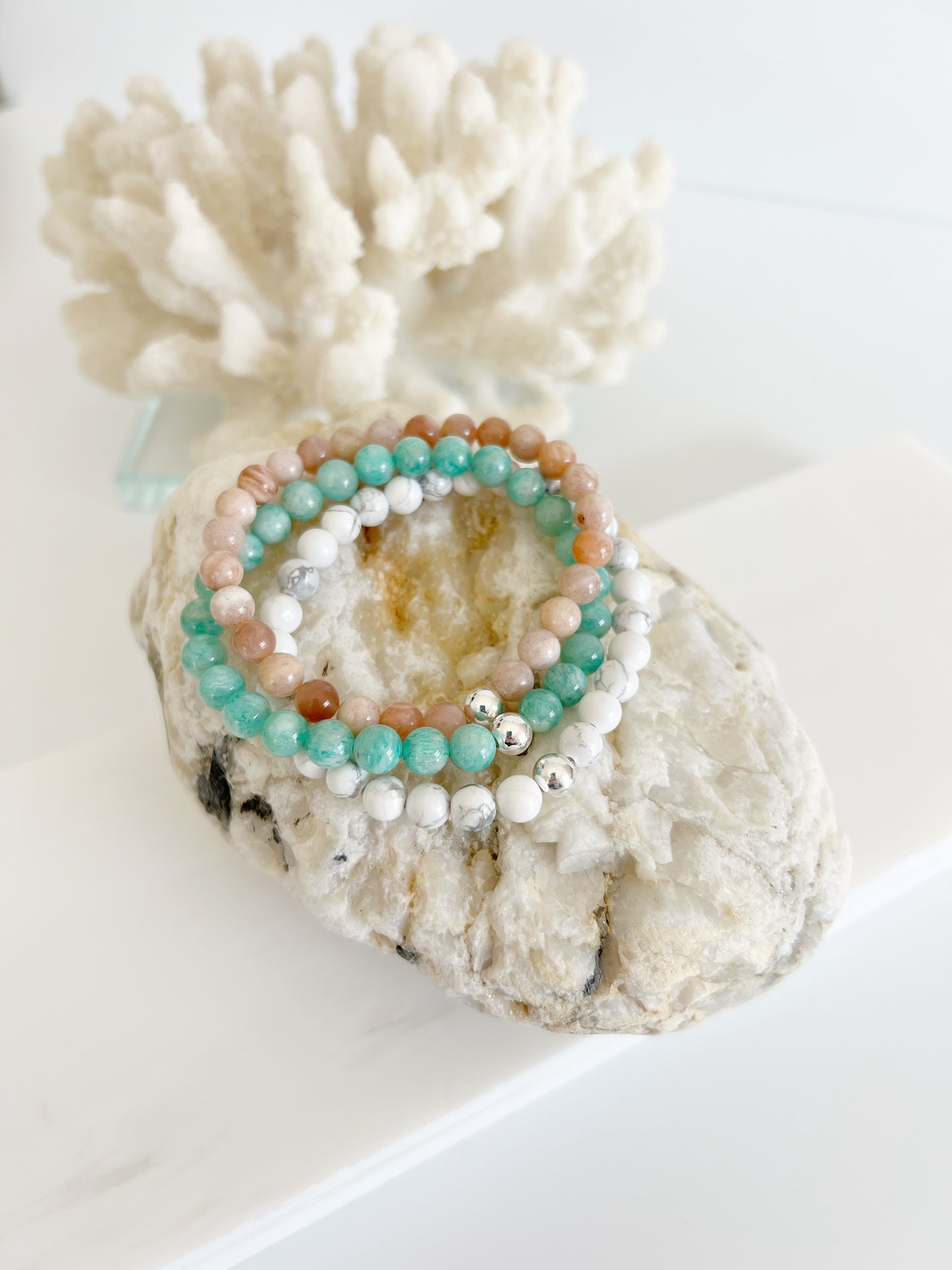 Three stretch bracelets stacked on top of each other, amazonite, peach moonstone and howlite with one silver bead on each on top of light brown rock with white coral behind it.