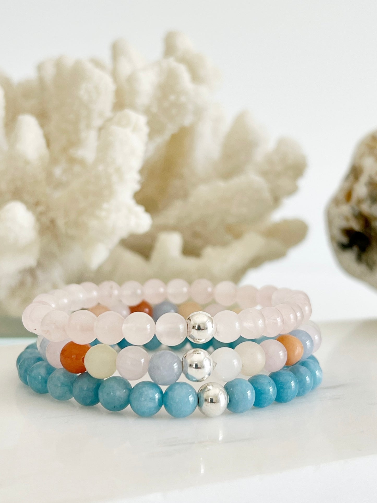 Three bracelet stack of stretch bracelets, a light baby pink one, a bright light blue and a multi colored one of light blue, light pink, yellow and deep orange in front of white coral and light brown rock.