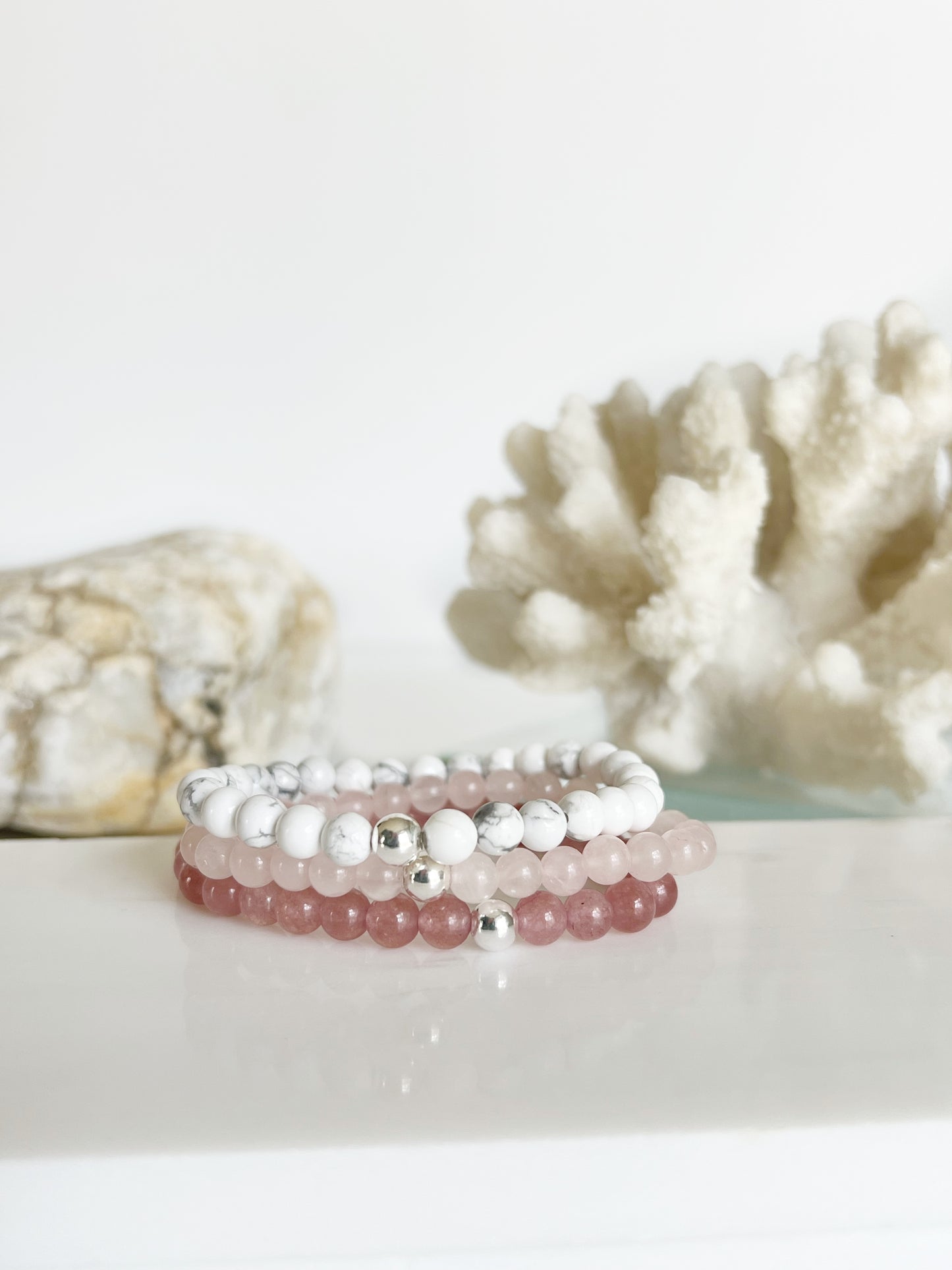 three stretch bracelets stacked on top of each other, a deep pink, light baby pink and white ones with one silver bead on each. They are in front of white coral and a light brown rock.