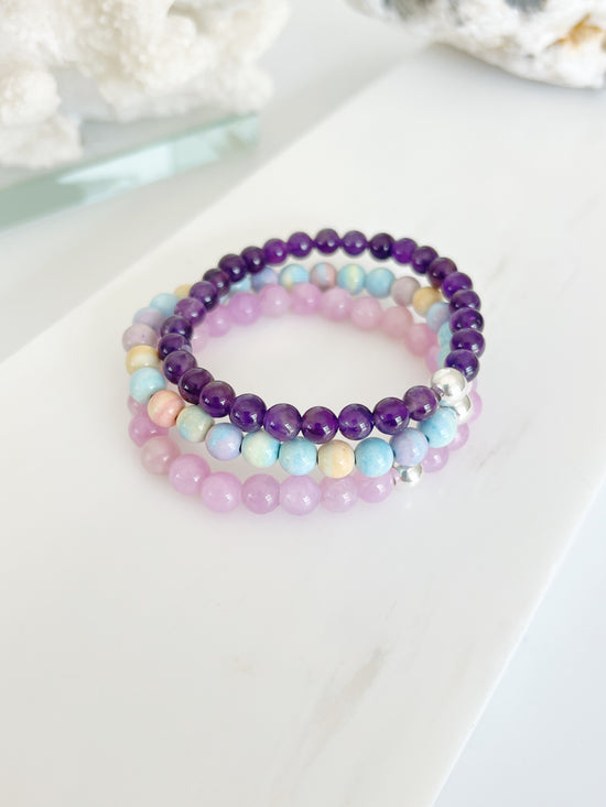 three stretch bracelets on top of each other, amethyst (deep purple ) a candy colored bracelet ( yellow, lilac, blue, green ) and a kunzite ( lilac ) 
