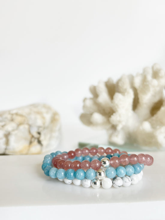 A bracelet stack of a white, bright light blue and deep pink bracelets stacked on top of each other. In front of white coral and a light brown rock