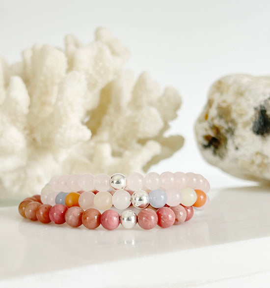 Three stretch bracelets stacked on top of each other, light baby pink, a multi colored one with pink, yellow, dark orange and blue, and an opaque dark pink one. Each one has a one silver bead. The are in front of white coral and light brown rock.