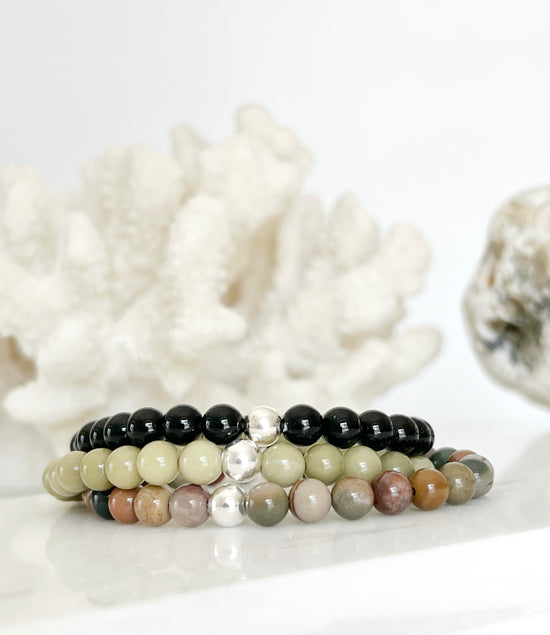 three stretch bracelets stacked on top of each other, black, avocado green and a multi green, red and brown one  with a silver bead each