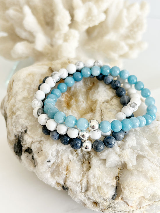 Three bracelets stacked on top of each other, dark blue, white and bright light blue with one silver bead on each. On top of a light brown rock with white coral behind it.
