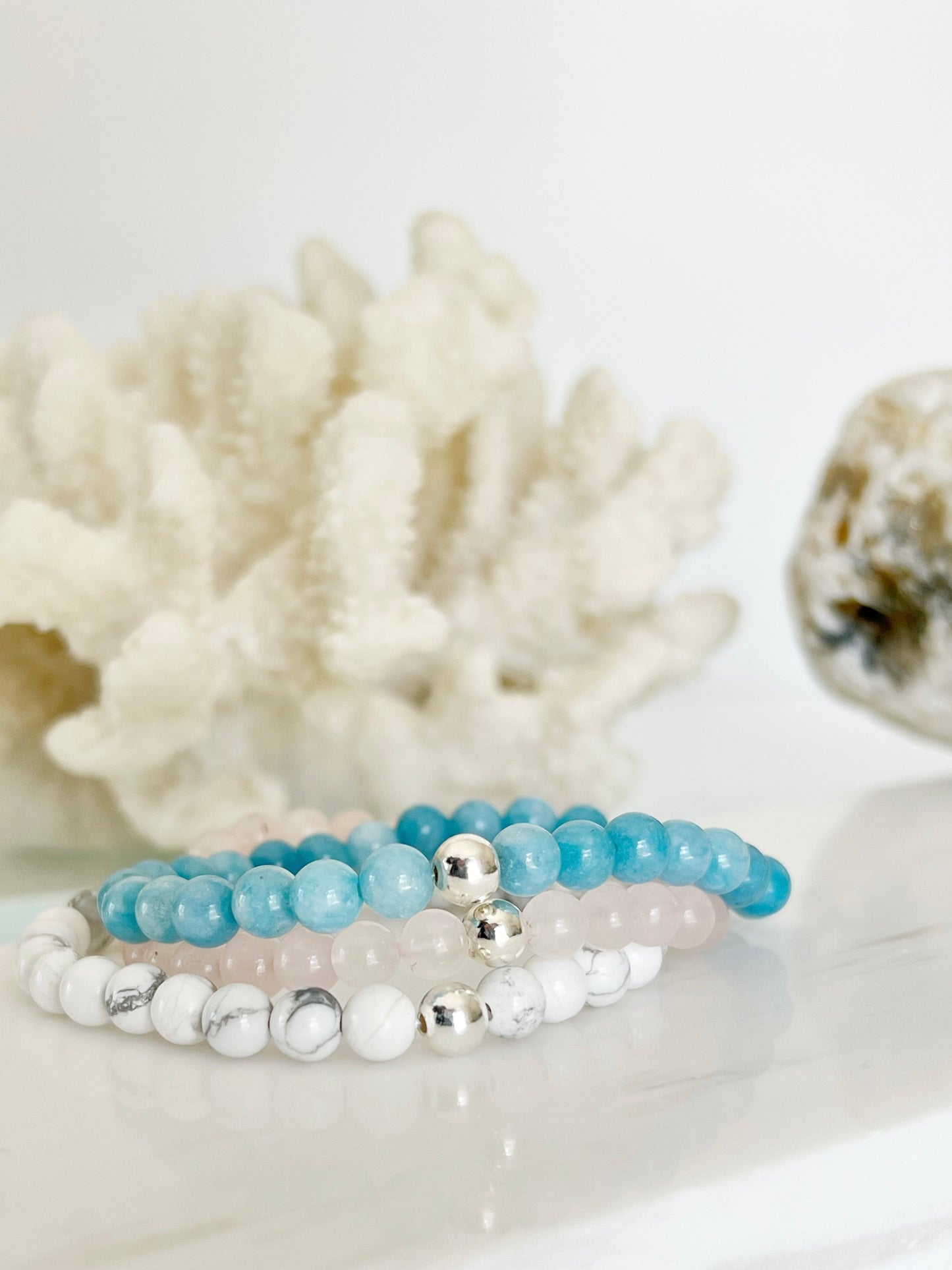 three stretch bracelets on top of each other in front of white coral with part of a rock on the right side. the bottom one is white, the middle is light pink and the top is light blue