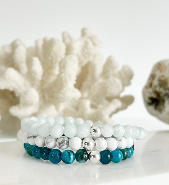 Three stretch bracelets stacked on top of each other. One is light green, one white and one mixed turquoise blue. They each have one silver bead and they are in front of white coral and a light brown rock.