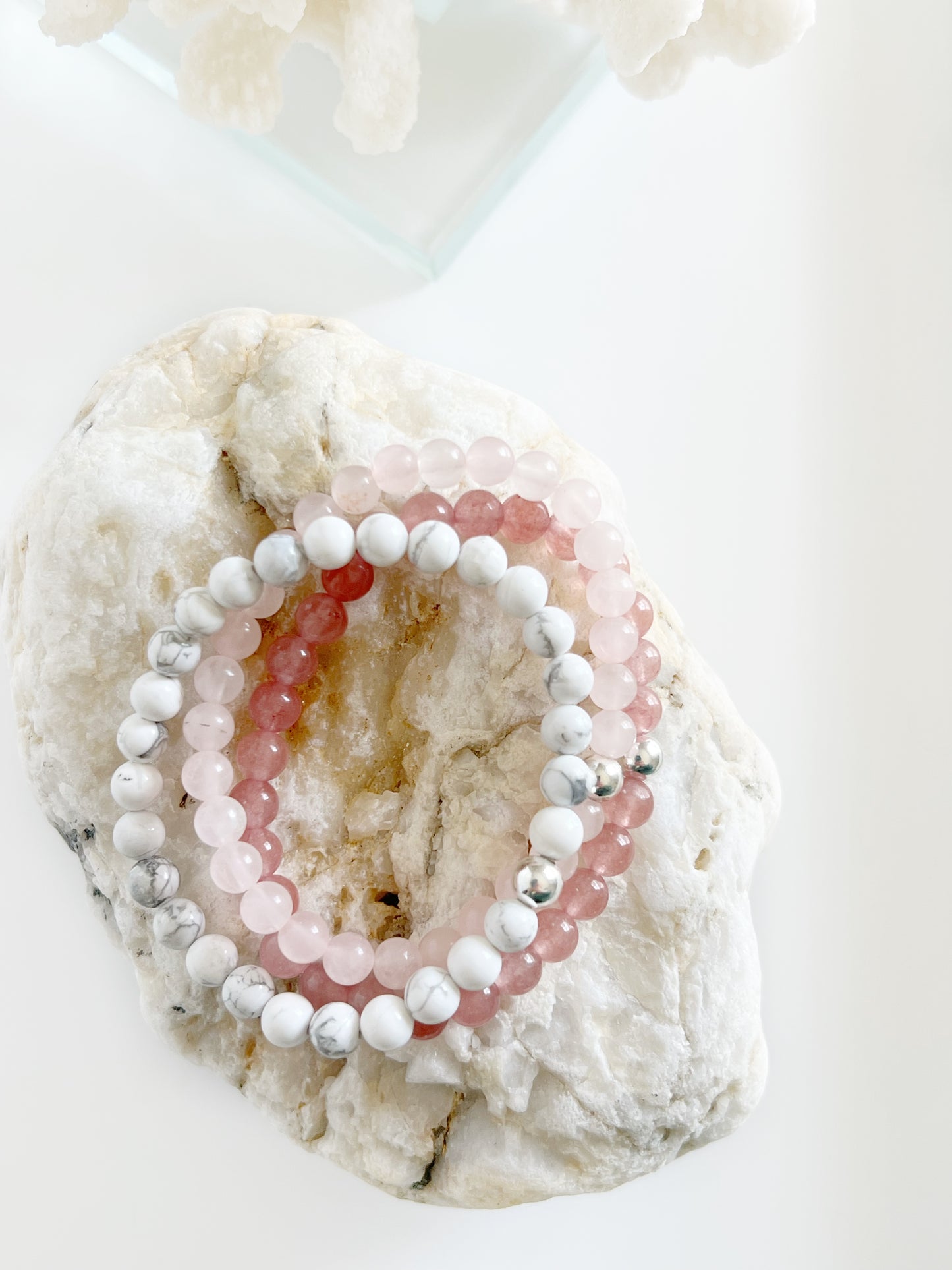 three stretch bracelets stacked on top of each other, a deep pink, light baby pink and white ones with one silver bead on each. All of top of a light brown rock.