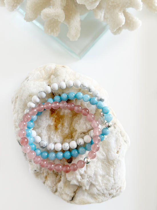 A bracelet stack of a white, bright light blue and deep pink bracelets stacked on top of each other sitting on top of light brown rock with white coral behind it.