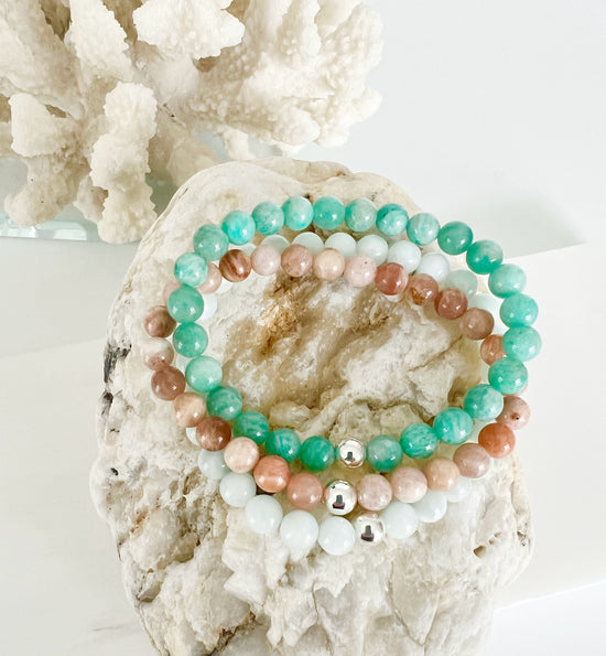 Simple everyday gemstone stretch bracelet trio with Amazonite, Peach Moonstone and Green Moonstone sitting on top of a brownish rock