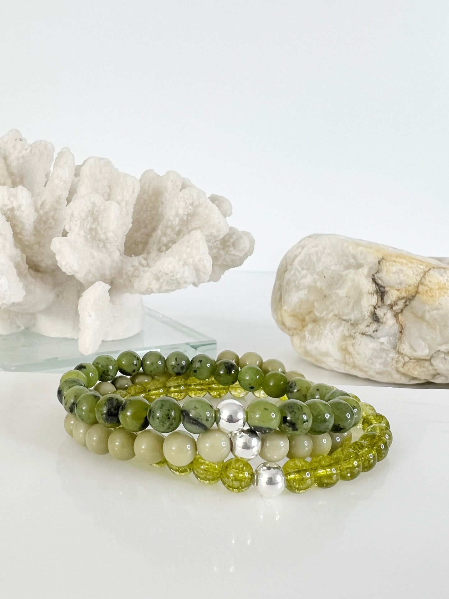 Green bracelet stack. 3 stretch bracelet, Canadian Jade, Avocado Jasper and Peridot. Each one with one sterling silver bead. There's a white coral and a brownish rock behind the stack. The bracelets are stacked on top of each other.