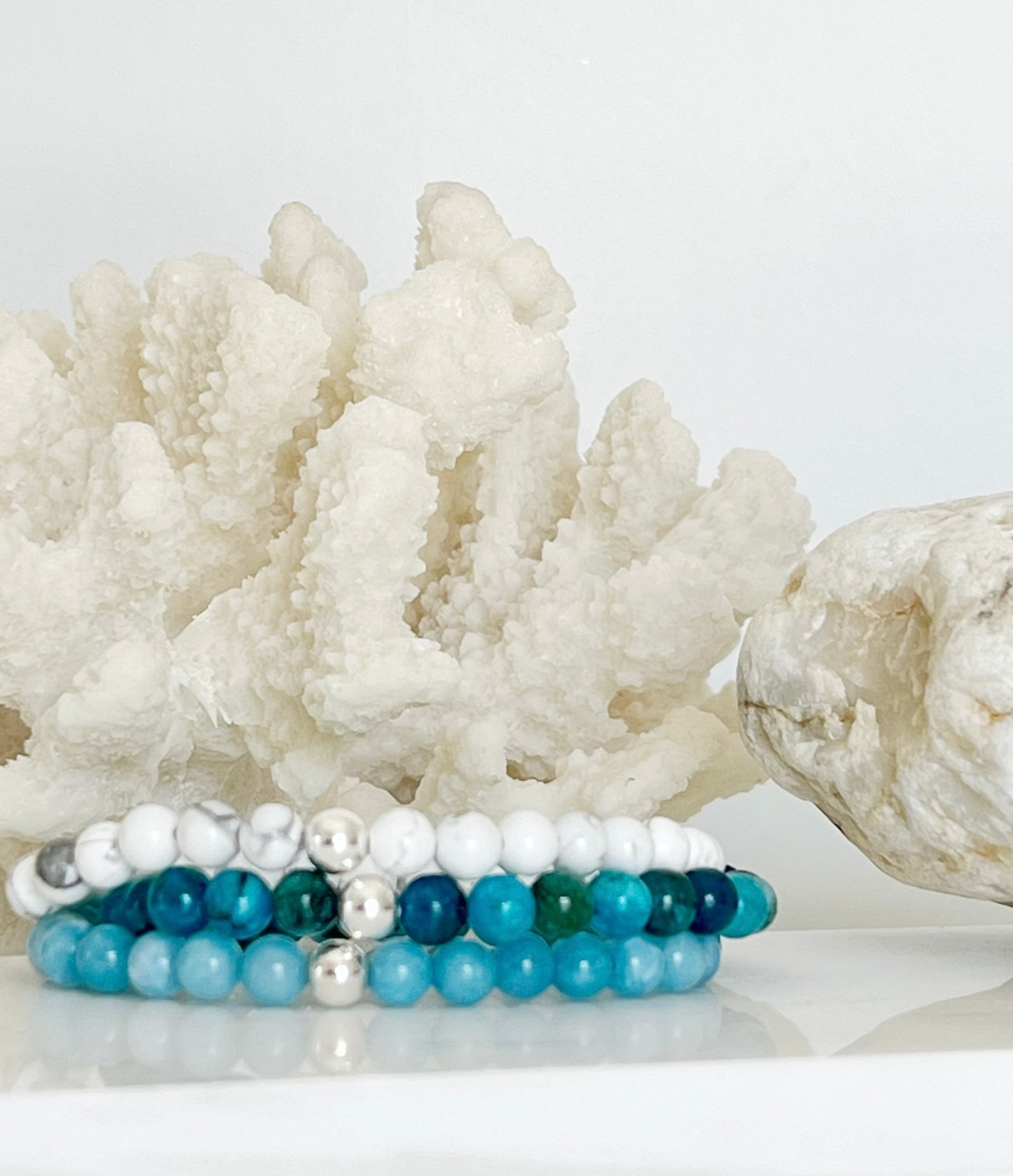 Three stretch bracelets stacked on top of each other in front of white coral and a brownish rock. The stretch bracelets are white, turquoise like ocean waves and light blue