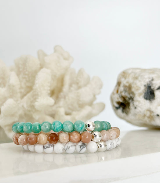 Three stretch bracelets stacked on top of each other, amazonite, peach moonstone and howlite with one silver bead on each. In front of white coral and a light brown rock.