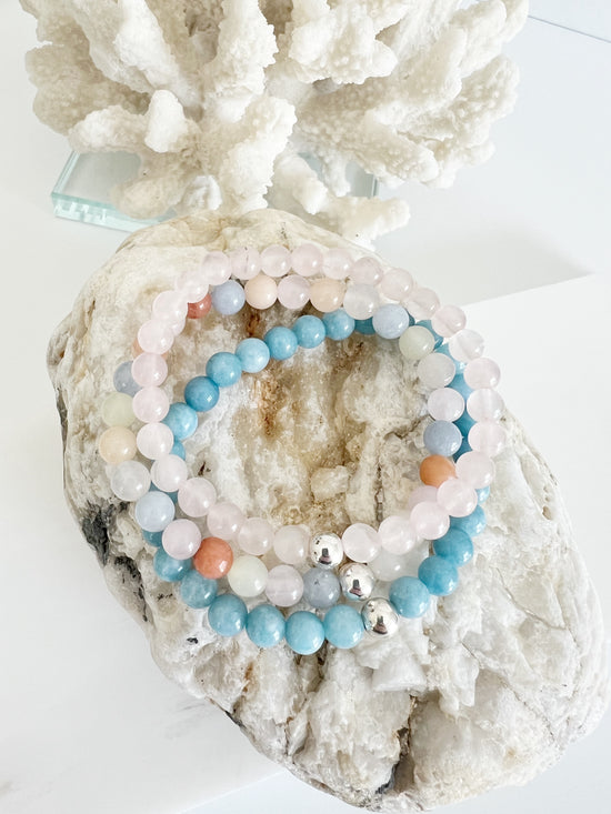 Three bracelet stack of stretch bracelets, a light baby pink one, a bright light blue and a multi colored one of light blue, light pink, yellow and deep orange on top of a light brown rock with white coral behind it.