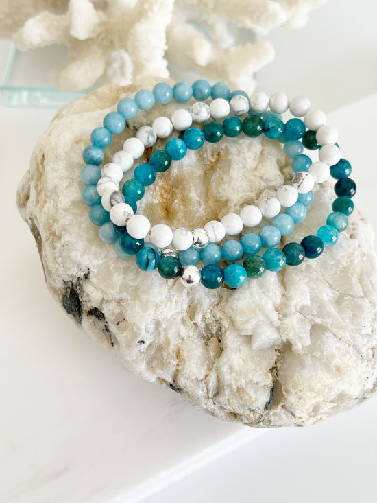 Three stretch bracelets, a white, a turquoise like the ocean and a light blue one on top of each other sitting on a brownish rock. Each bracelet has one silver bead on it.