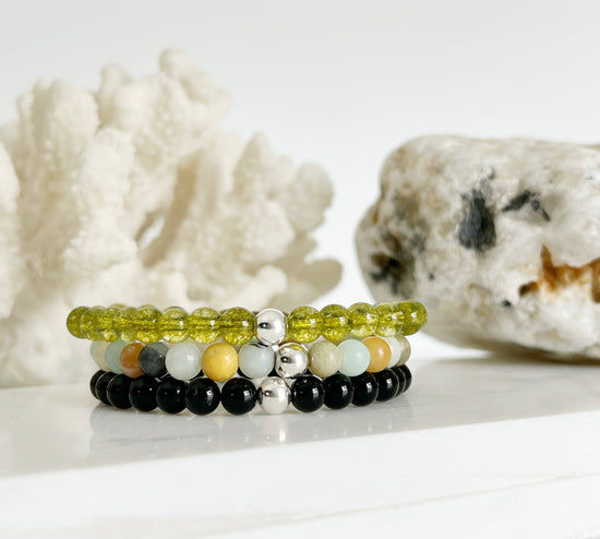 three stretch bracelets stacked on top of each other in front of white coral and light brown rock. One transluscent green one, a multi colored one with light blue, yellow, black and white and a black one. All have one silver bead.