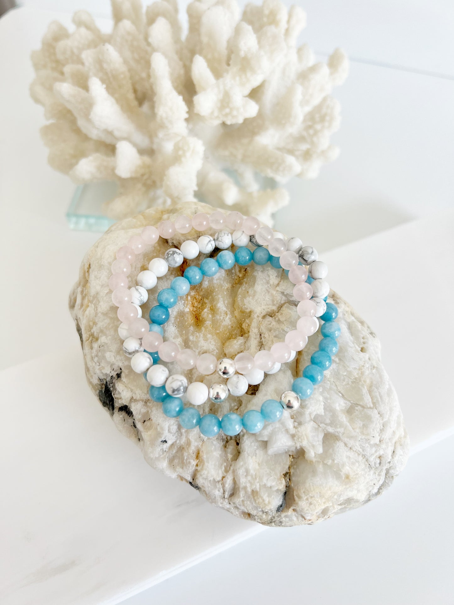 three bracelets on top of a whitish brown rock with white coral behind it. the top bracelet is light pink the middle is white and the bottom is light blue