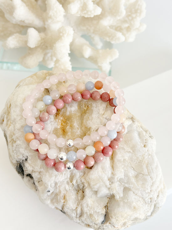 Three stretch bracelets stacked on top of each other, light baby pink, a multi colored one with pink, yellow, dark orange and blue, and an opaque dark pink one. Each one has a one silver bead on top of a light brown rock in front of white coral.
