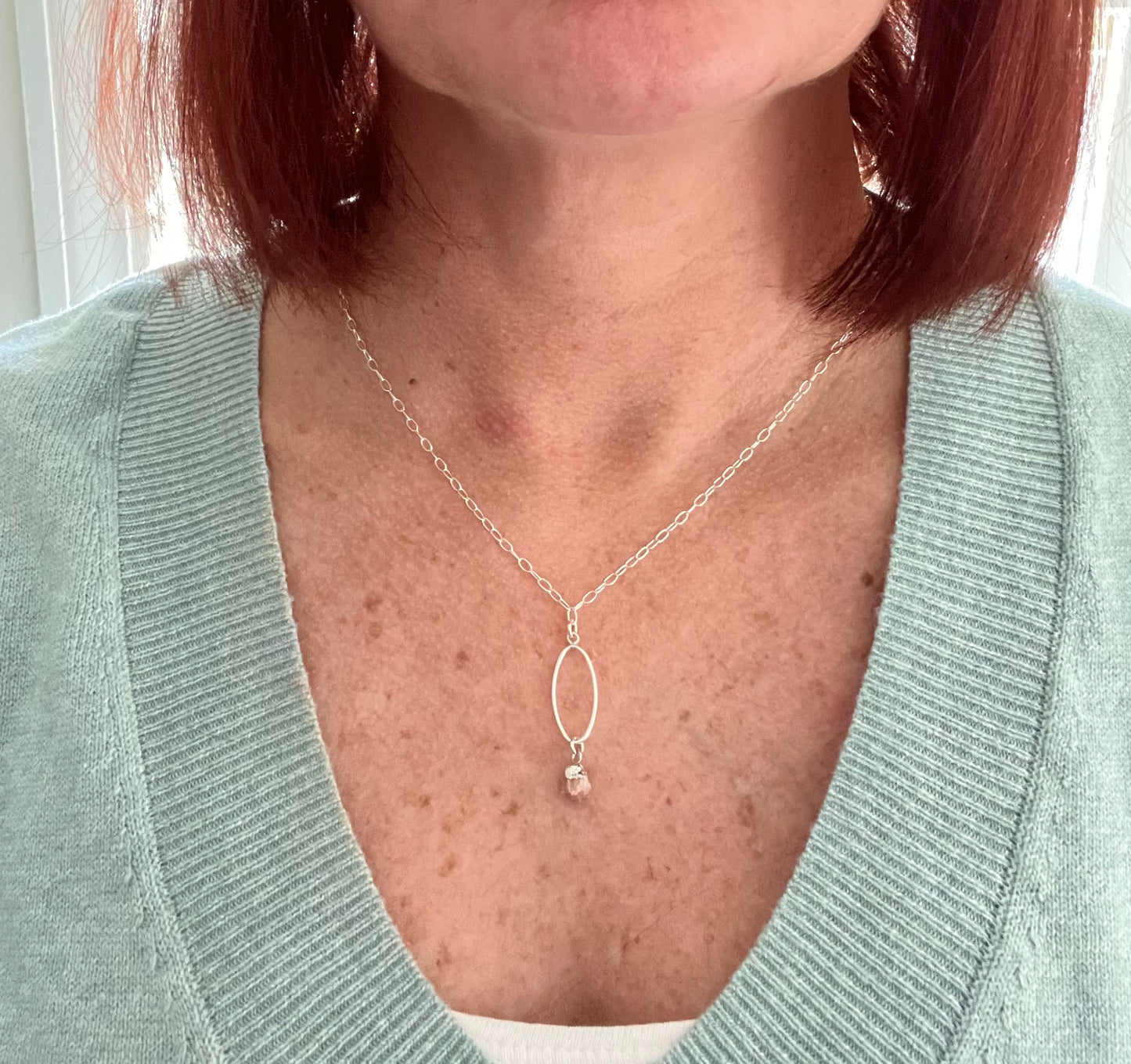 A woman wearing a sage green v neck sweater with a white tank top underneath. Her necklace is sterling silver and the pendant is a sterling long flat oval with a pink tourmaline bead hanging on the bottom.