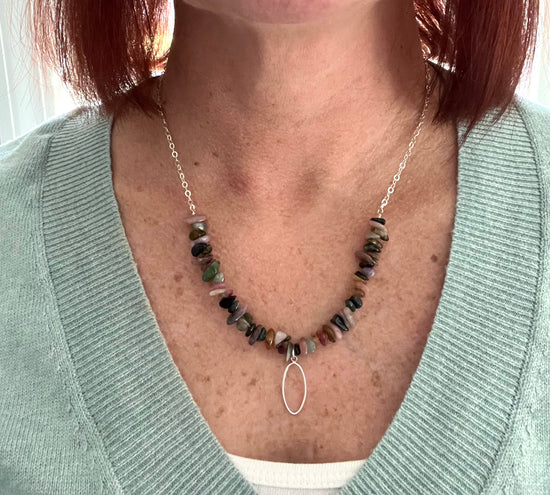 a woman wearing a sage green v neck sweater and a white tank top underneath. Her necklace is a sterling silver chain with tourmaline chips and a dangling silver flat oval on the bottom.