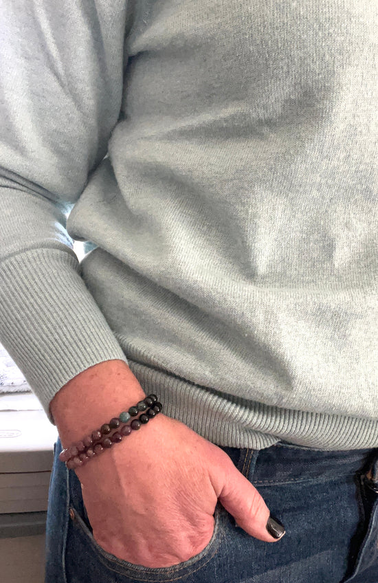 A picture showing a woman's torso with her hand in her pocket. She's wearing a sage green sweater and jeans and she has two dark Tourmaline bead stretch bracelets on.